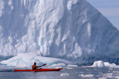 Eco tourist in single sea kayak paddles by an iceberg, watched by a penguin. Brown Bluff. Antarctica