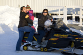 Holding a child and a bottle of 'Coke' a young Inuit woman climbs onto the back of a snowmobile outside the Co-op in Pangnirtung. Nunavut, Canada. 2008