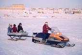 An Inuit girl drives a snowmobile towing other Inuit children on a sled. Baker Lake, Nunavut, Canada. 1982