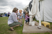 Mother watches her children try to milk a fake cow. Sturminster Newton Cheese Festival. Dorset. England
