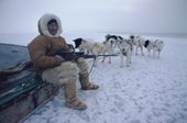 Fur clad Inuit hunter, Avataq, sitting on his sled with his rifle, and dog team behind. NW Greenland. 1980