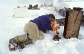 Inuit hunter, Qaavigannguaq lights a Primus stove to make tea during a rest on a Spring seal hunt. He wears polar bear pants & sealskin kamik (boots). Moriussaq, Northwest Greenland. 1980