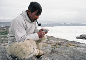Angutikavsaq, an Inuit man, smokes his pipe whilst carving a small soapstone figure. He is wearing polar bear skin trousers and seal skin kamik (boots). Qeqertat, Northwest Greenland. (1980)